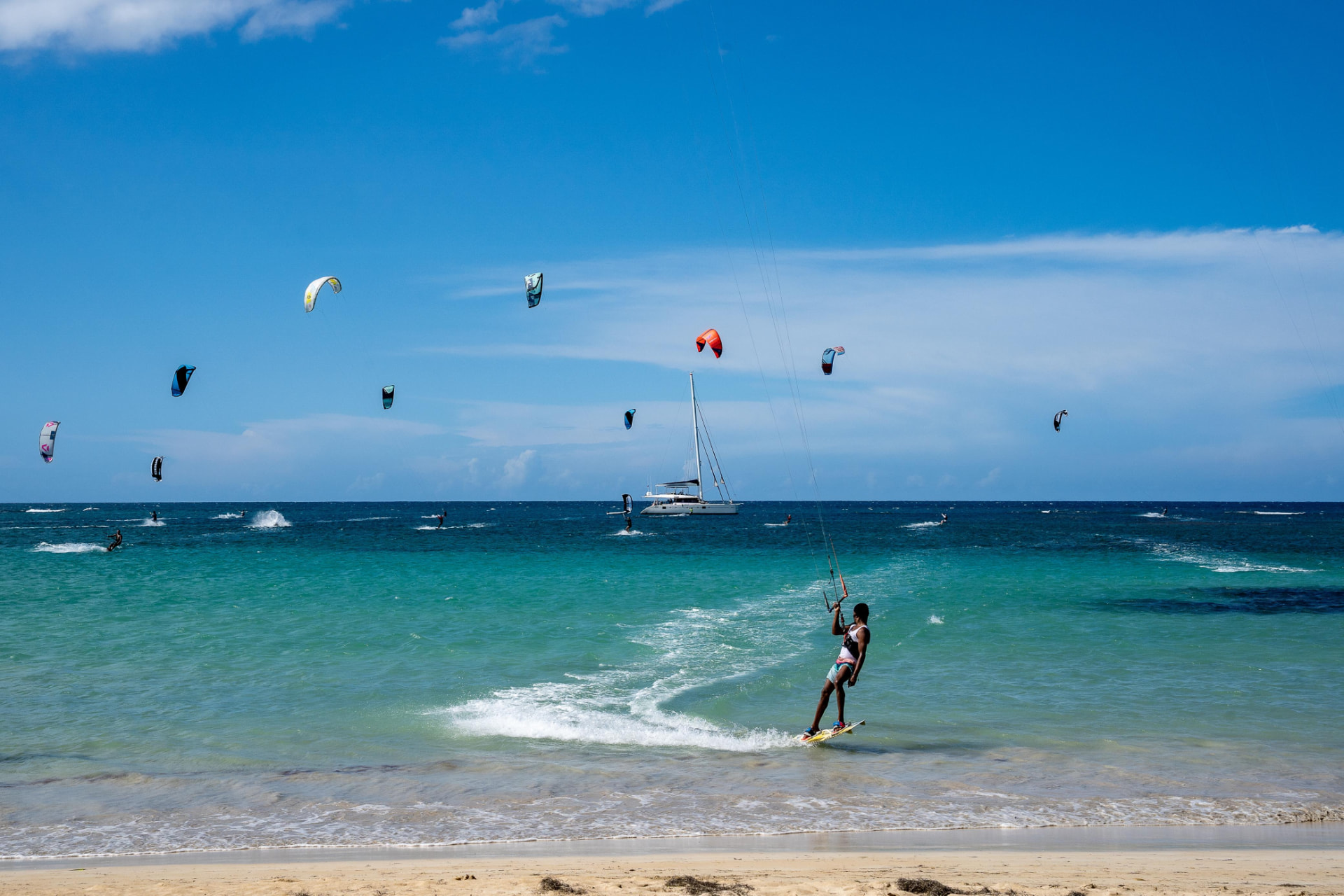 A bright turquoise ocean with kite surfers and a sailboat under a clear blue sky